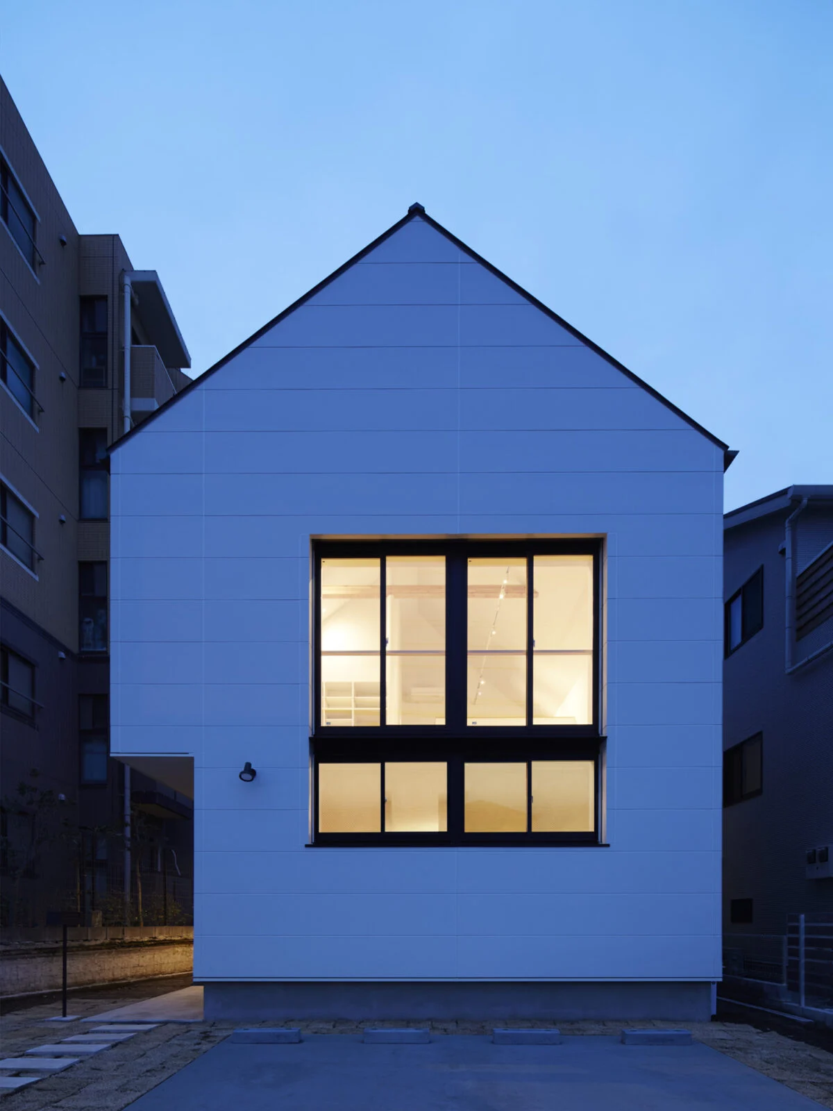 外観　夕景／三角屋根に白い壁。子どもが描く「おうちの家」そのままのような、愛らしくてホッとする外観。上下階で開口の幅をそろえてあり、まるで1つの大きな窓のよう。2階のLDKを広くしたため、1階の左が少し凹んでいるが、屋根の重みを家の中心に流す設計で外柱が不要になり、シンプルでかわいらしいシルエットが実現した。ここには玄関があるので雨の日の庇（ひさし）がわりにもなるそう