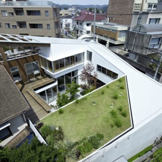 中庭を中心に住空間をスパイラル状に構成。手前がピアノ教室の屋上につくられた屋上庭園、左の茶色の屋根が既存住宅。奥は1階がLDK。デッキテラス奥の木製外壁部分は中2階の浴室と屋上テラス。その右の白壁部分は2階の寝室。道路側は1階がメインエントランス、中2階が書斎