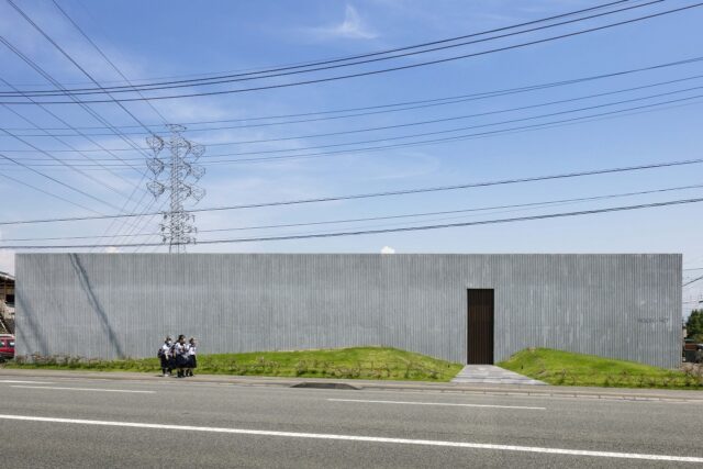 40mの壁が印象的なファサード。甲府市内の幹線道路沿いという都市スケールにマッチする、堂々たる佇まいだ。陰影のある出目地コンクリートと芝のマウンドによるシンプルな構成は、幹線道路の無機質な景色にうるおいをプラス。公園のような存在感がある