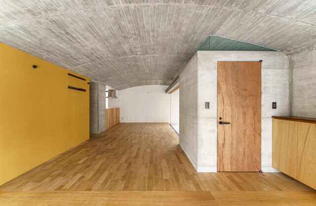 １階、玄関から生活空間を見る。右の扉はトイレへ続く。緩やかなアールを描く天井を生かすため、天井には照明器具を取り付けなかった。画像右奥のテラスの外へと天井は続く。左側壁面の親しみが感じられる黄色は、沖縄の土由来のものだという

