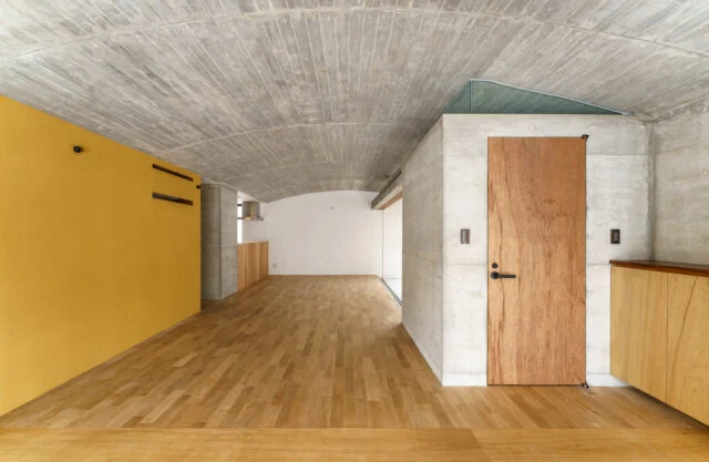 １階、玄関から生活空間を見る。右の扉はトイレへ続く。緩やかなアールを描く天井を生かすため、天井には照明器具を取り付けなかった。画像右奥のテラスの外へと天井は続く。左側壁面の親しみが感じられる黄色は、沖縄の土由来のものだという

