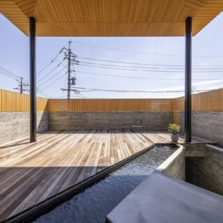 玄関脇からテラスを見る。大屋根に覆われ半屋外的な雰囲気だ。画像右の空間を下ると車庫がある。要望もあり水盤はダイナミックに配置した。水紋や日に反射する水の表情を室内からも楽しめる。テラスの壁は木板を裏表でずらして計画。視線は遮り、風は通す