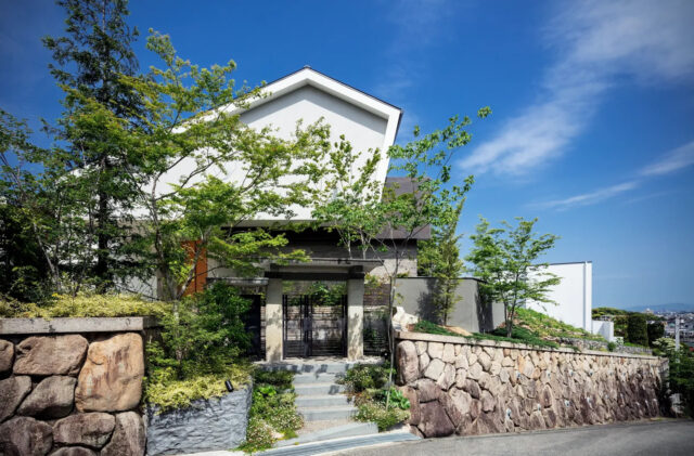 外観。家を建て替えるにあたり、鬱蒼としていた木々を整備するなど、まずは土地の整備から始めた。既存の石垣や画像中央の門扉などシンボル的なものは補強をしながら残したという。Sさまのさりげなくという意を汲み取り、家が木々で隠れるように計画
