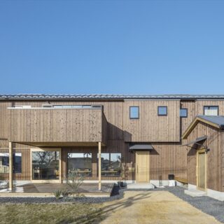 外観。道路に接する面がとても長い敷地の特徴を活かし、建物もあえて横長に。右手前の小屋は施主様の事務所。横長で大きく見える建物と、かわいらしい小屋の対比が印象的だ。特に意識したのが、2階のデザイン。建物が大きいので、外壁が平面だと圧迫感が出る。そのため、飛び出した部分を何か所か設けて変化をつけている
