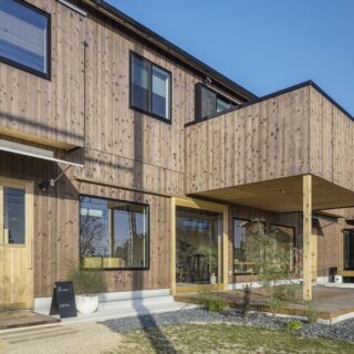 前庭から見た建物。1階がカフェで2階が住居スペースとなる。2階には広いベランダが設けられ、その下はカフェのテラス席として使用されている。1階のカフェには大きな窓を設置し、室内の明るさを確保している。また、人の手が触れる部分には木製の明るい部材を使用し、暖かみを感じられるように計算されている
