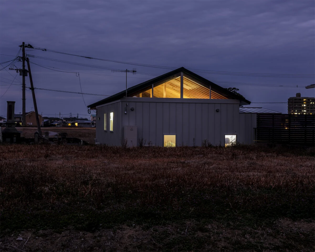 住宅街側から見た夕景。現在は空き地となっている隣地に、できる限り寄せて建てられている。下に寄せて窓を設けることで、窓を開けても室内が見えないように配慮されている。その窓はすりガラスで室内は見えないが、ハイサイドライトは透明で美しい梁がよく見える
