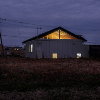 住宅街側から見た夕景。現在は空き地となっている隣地に、できる限り寄せて建てられている。下に寄せて窓を設けることで、窓を開けても室内が見えないように配慮されている。その窓はすりガラスで室内は見えないが、ハイサイドライトは透明で美しい梁がよく見える
