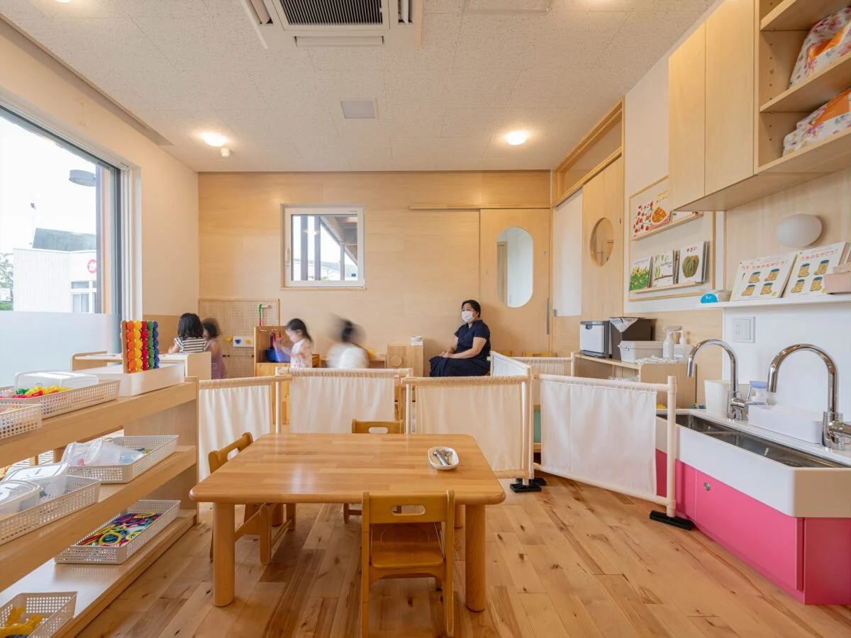 1歳児室。右奥、正面の扉はサンルーム、0歳児室へ続く。右の扉はトイレ。部屋はL字で水道の脇から右へ伸びる。そのエリアを前室とし、室内で遊ぶ園児たちから届け物などをしに来た親の顔が見えないように工夫した。園児の目線の高さでフロストシートを貼り外からの視線を遮った