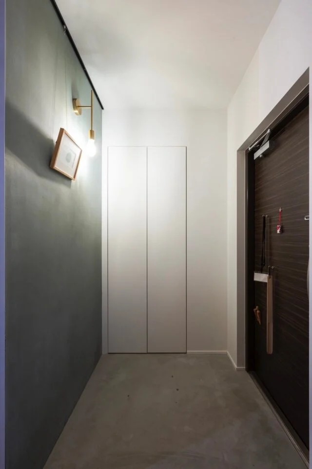 玄関。入口正面はブルーグレーの漆喰壁を背景としたピクチャーレール付きウォールで、真鍮ブラケット照明と共に住宅の第一印象を彩る