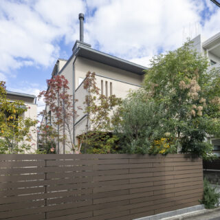 外観の形は違えど、屋根の勾配やベージュの外壁の色が揃った２棟。板塀でつながり、中では庭を共有している。板塀の上から顔を覗かせるさまざまな樹々は、まるで訪れる人を出迎えているかのようだ