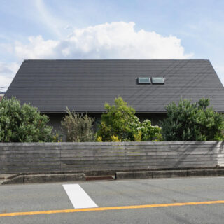 敷地の北を走る幹線道路から見た「サンカクヤネノイエ」。車の騒音や排気ガスから生活空間を守るために大屋根と木塀で覆い、1階の窓部分は植栽で目隠ししている。2階はほとんど窓がなく全体的に閉じた印象だが、邸内にはこの外観から想像もつかない明るい大空間が広がる
