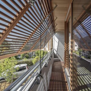 2階テラス。上から３分の１あたりに回転軸をもつ木製格子戸。紐を引き、跳ね上げ固定する。右は広間に続く木製サッシ。軒の深さと格子戸で雨掛かりのリスクが低くなることからアルミではなく木製の、さらに珍しい国産スギのサッシを採用した