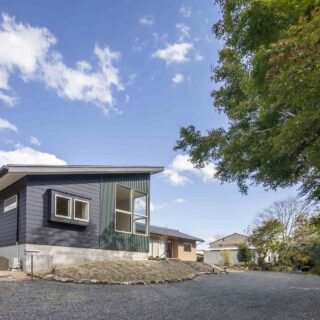 道路側から見た外観。子世帯の外壁は緑あふれる竹林などの自然に調和する、グリーンとブラック。手前の砂利部分が車の通行・駐車エリアだ。高低差を活用した配置のため、道路からの視線もカットされている。奥の親世帯は家の向きと子世帯の建物の影で視線をカットしている