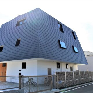 白い軸の上に、グレーの傘がのった「キノコ型」のH邸。１階の壁は長尺のサイディングに塗装し漆喰のような風合いを持たせた。２－３階の壁と屋根は、ガルバリウム鋼板を採用。