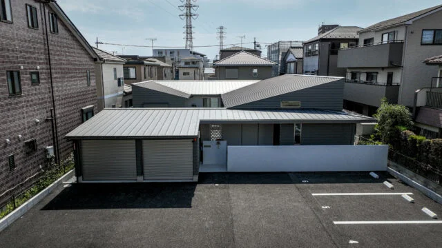 三方を3階建て住宅に囲まれた敷地に建つ平屋。手前は貸し駐車場として使われており、不特定多数の目が届くため、建物は敷地の奥へと配された。