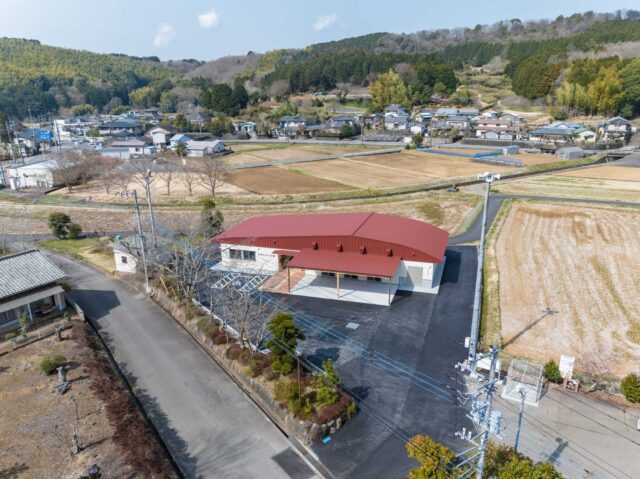 周りを山々に囲まれた山間の集落に建つ田原野公民館（東側からの外観全景）。約50世帯が集まる集落の中心部に位置し、田畑に囲まれたのどかな環境が広がる