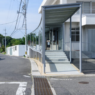 道路と敷地の形状にあわせ、自然な形で配置された増築部。曲線や勾配がある近隣道路の特徴を、そのまま取り込んだ。無理やり造形しているわけではない。写真右奥には新設したスロープが見える。駐車場後ろのスペースを有効活用し、駐車台数を減らしていない点に工夫の跡が見られる
