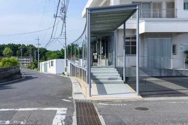 道路と敷地の形状にあわせ、自然な形で配置された増築部。曲線や勾配がある近隣道路の特徴を、そのまま取り込んだ。無理やり造形しているわけではない。写真右奥には新設したスロープが見える。駐車場後ろのスペースを有効活用し、駐車台数を減らしていない点に工夫の跡が見られる