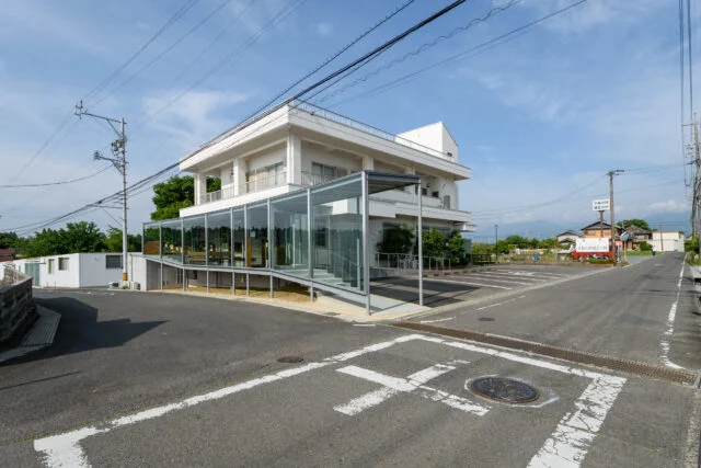 外観。敷地の角まで増築部があるため、受け壁もガラスとした。写真左の道から来た車が写真右側へ左折する際、人や車の交通状況がわかるようにと配慮した結果だ。全面ガラスの側面との連続性をあわせ持つ、違和感のないデザインとなっている