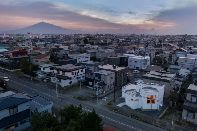 上空から。津軽富士と呼ばれる岩木山を遠方に望む住宅地。独創的な外観が目を引く。外壁が白い影響で建物が大きく見えるため、屋根を低く抑えていることがわかる